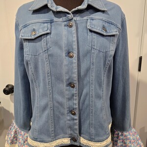 May include: Light blue denim jacket with a classic collar, button-up front, and two chest pockets. The cuffs feature a floral print with a lace trim. The jacket is displayed on a mannequin.