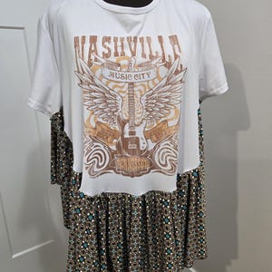 May include: A white short-sleeve t-shirt with a raw-edge hem featuring a brown graphic of a guitar, wings, and the text Nashville Music City Rock Star Tennessee. The lower portion of the garment is a gathered skirt with a brown, black, and teal pattern.