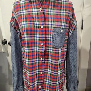 Op de afbeelding: Een overhemd met knopen met een rood, blauw, groen en wit geruit patroon op de voor- en achterkant. De mouwen zijn van blauwe denim met kleine witte stippen. Het overhemd heeft een enkele borstzak.