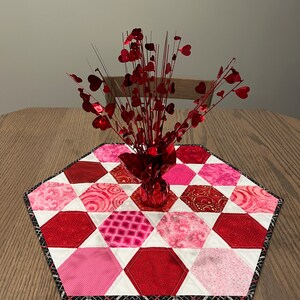 May include: A red and white hexagonal table runner with a red heart-shaped floral centerpiece. The runner is made of fabric with different patterns and is edged with black trim.