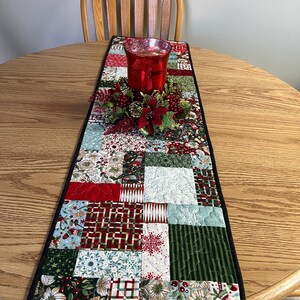 May include: A quilted table runner with a patchwork design featuring red, green, white, and blue fabrics. The runner is trimmed with black fabric and has a festive Christmas theme.