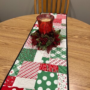 May include: A red and green Christmas table runner with a black border. The runner is made of patchwork fabric with various holiday patterns, including snowmen, candy canes, and snowflakes. A red glass candle holder with a green and red floral arrangement sits on the runner.