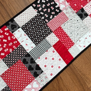 May include: A black and red patchwork table runner with a variety of patterns, including hearts, polka dots, and geometric designs. The runner has a black border.