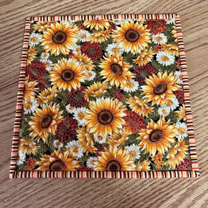 May include: A square fabric placemat with a black background and a pattern of yellow sunflowers, white daisies, and red and orange flowers. The placemat has a brown, orange, and white striped border.