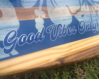 May include: Wooden sign with a blue background and the words "Good Vibes Only" in white script. The sign has a floral design and a natural wood border. The sign is resting on grass.