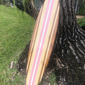 Puede incluir: Una tabla de surf de madera con rayas rosas. La tabla de surf est&aacute; apoyada en un tronco de &aacute;rbol.