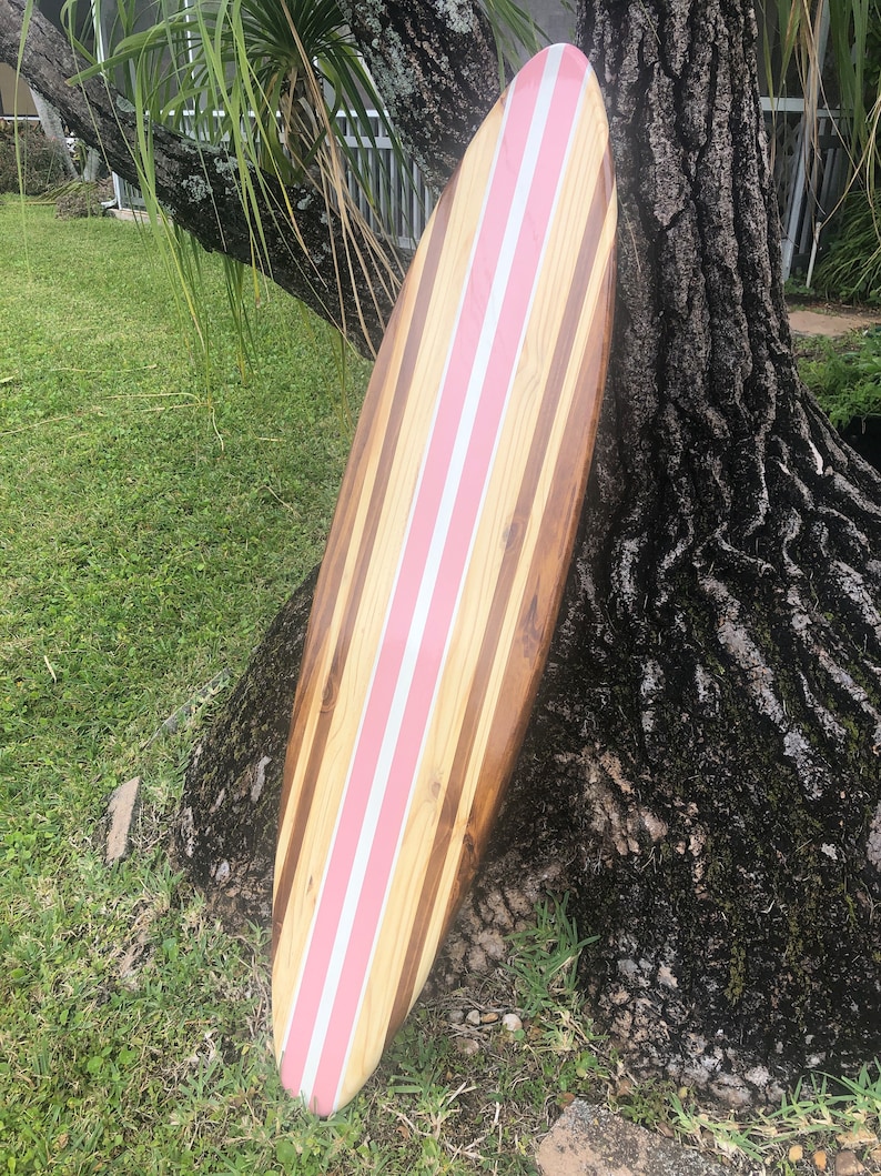 Puede incluir: Una tabla de surf de madera con rayas rosas. La tabla de surf est&aacute; apoyada en un tronco de &aacute;rbol.