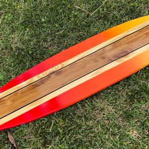 May include: A long, wooden surfboard painted in a gradient of yellow, orange, and red. The surfboard has three black stripes running down the center.