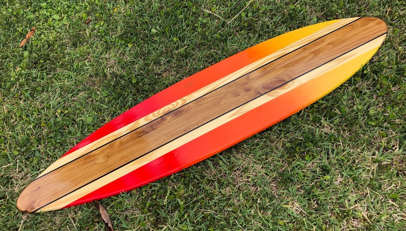 May include: A long, wooden surfboard painted in a gradient of yellow, orange, and red. The surfboard has three black stripes running down the center.