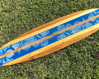 Puede incluir: Una tabla de surf de madera con un diseño floral azul y blanco. La tabla de surf está pintada con una franja azul y flores de hibisco blancas.