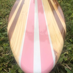 Puede incluir: Una tabla de surf de madera con rayas rosas y blancas. La tabla de surf est&aacute; hecha de diferentes tipos de madera, incluyendo marr&oacute;n claro, marr&oacute;n oscuro y madera marr&oacute;n rojiza.