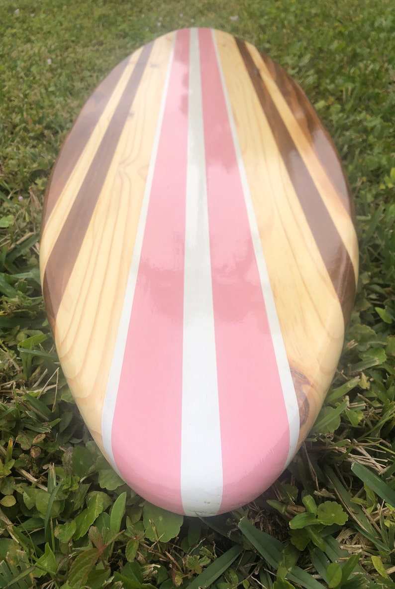 Puede incluir: Una tabla de surf de madera con rayas rosas y blancas. La tabla de surf est&aacute; hecha de diferentes tipos de madera, incluyendo marr&oacute;n claro, marr&oacute;n oscuro y madera marr&oacute;n rojiza.