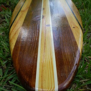 Op de afbeelding: Een houten surfboard met een natuurlijke houten afwerking en zwarte en witte strepen. Het surfboard is gemaakt van verschillende soorten hout, waardoor een uniek patroon ontstaat.