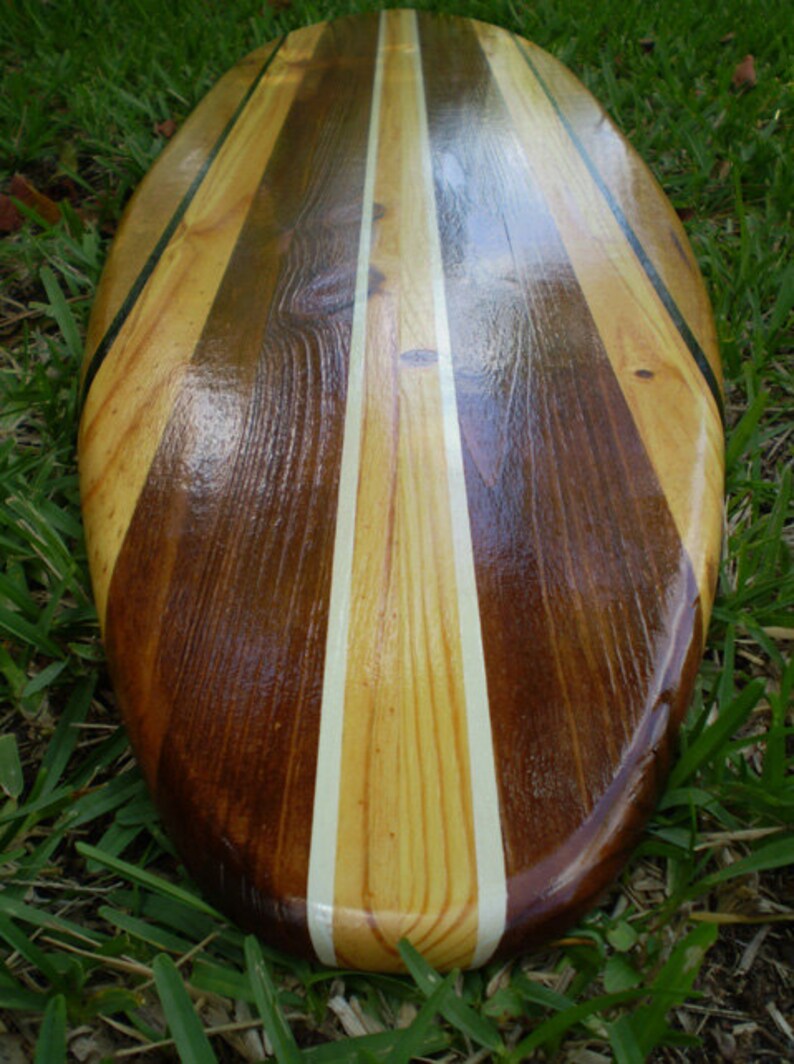 Op de afbeelding: Een houten surfboard met een natuurlijke houten afwerking en zwarte en witte strepen. Het surfboard is gemaakt van verschillende soorten hout, waardoor een uniek patroon ontstaat.
