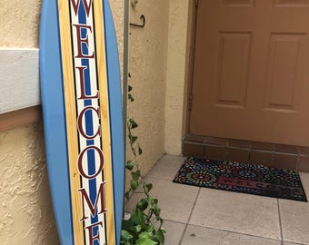 May include: A blue and white surfboard with a wood grain design and the word "WELCOME" written vertically in red letters.