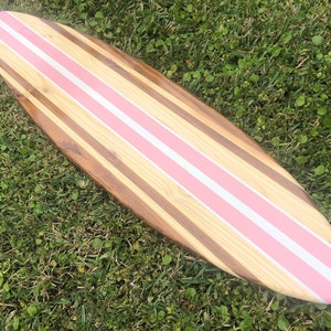 Puede incluir: Una tabla de surf de madera con rayas rosas y blancas pintadas en la parte superior. La tabla de surf est&aacute; hecha de diferentes tipos de madera, creando un patr&oacute;n &uacute;nico.