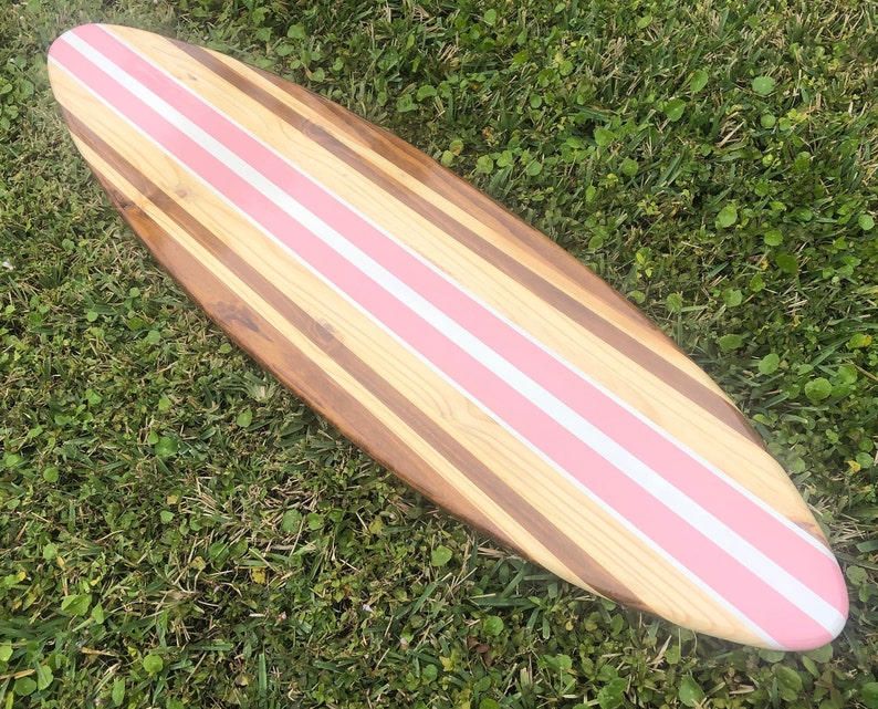 Puede incluir: Una tabla de surf de madera con rayas rosas y blancas pintadas en la parte superior. La tabla de surf est&aacute; hecha de diferentes tipos de madera, creando un patr&oacute;n &uacute;nico.