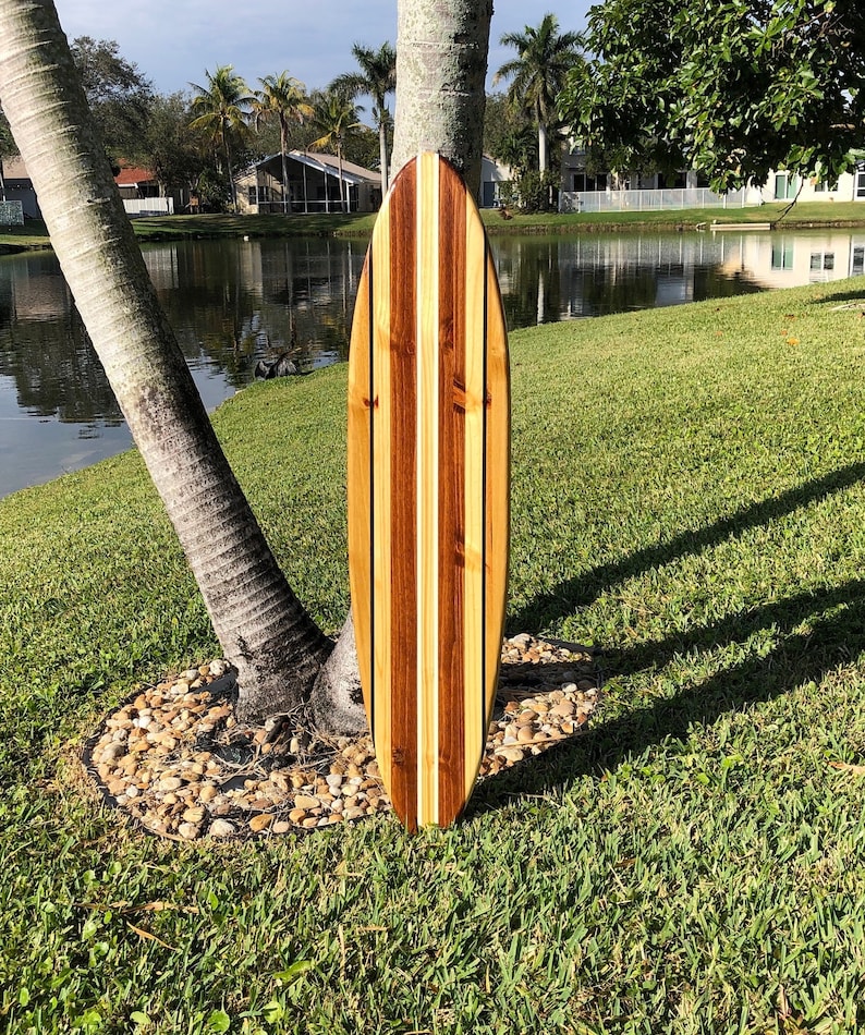 Op de afbeelding: Een houten surfboard met een gestreept ontwerp in lichtbruin, donkerbruin en wit. Het surfboard leunt tegen een boomstam in een grasachtig gebied.