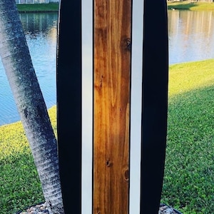 May include: A wooden surfboard with a glossy finish. The board features a central panel of natural wood, flanked by white stripes and black edges. The board is approximately 6 feet long.