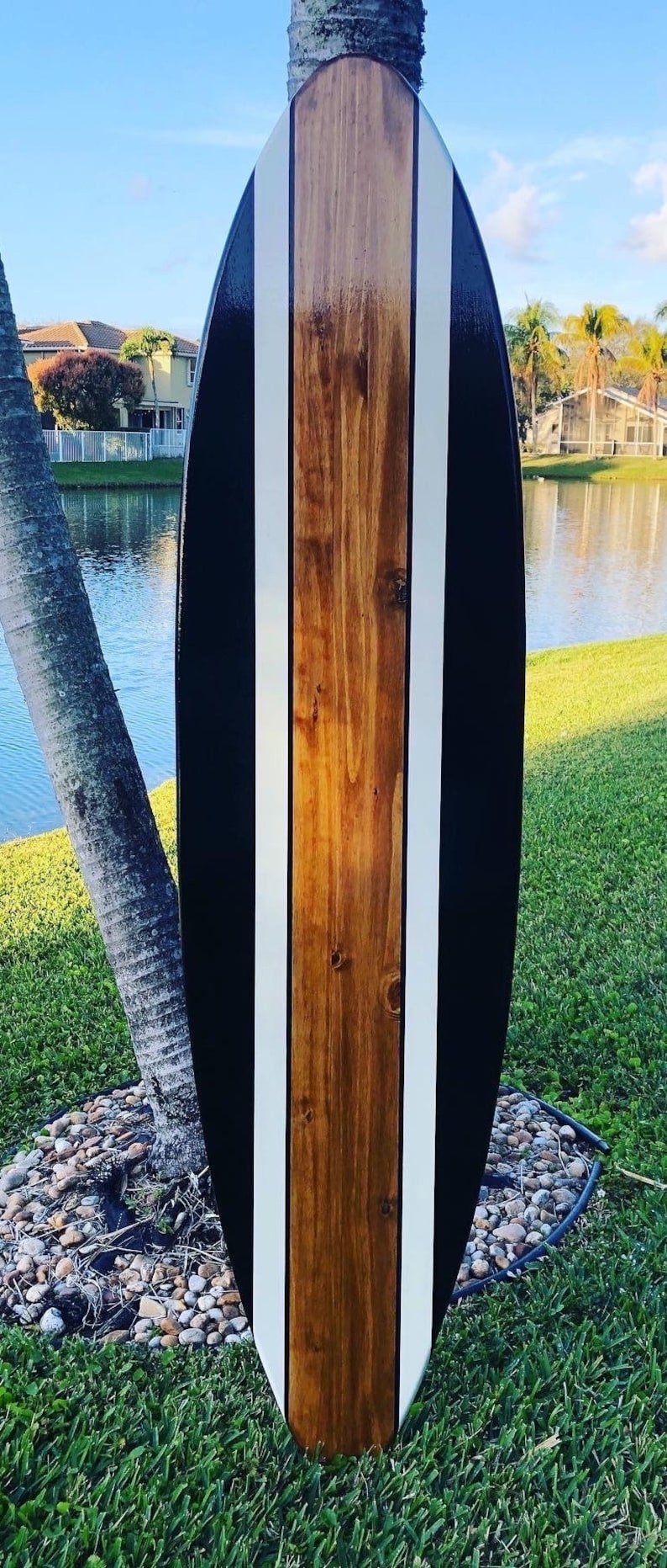 May include: A wooden surfboard with a glossy finish. The board features a central panel of natural wood, flanked by white stripes and black edges. The board is approximately 6 feet long.