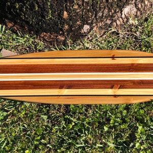 Op de afbeelding: Een houten surfboard met een natuurlijke houten afwerking en twee witte strepen. Het surfboard is gemaakt van verschillende soorten hout, waaronder ceder en walnoot.