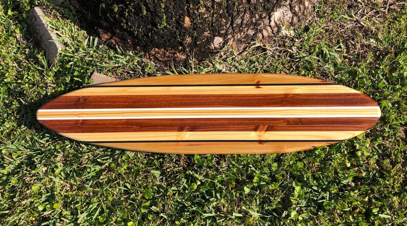 Op de afbeelding: Een houten surfboard met een natuurlijke houten afwerking en twee witte strepen. Het surfboard is gemaakt van verschillende soorten hout, waaronder ceder en walnoot.