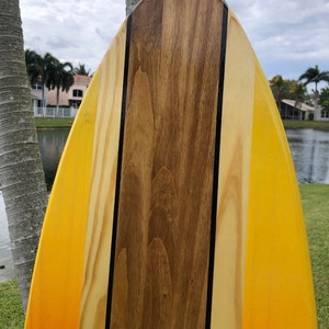 May include: A wooden surfboard with a yellow and orange gradient paint job. The surfboard has a black stripe down the center and a brown wood panel in the middle.