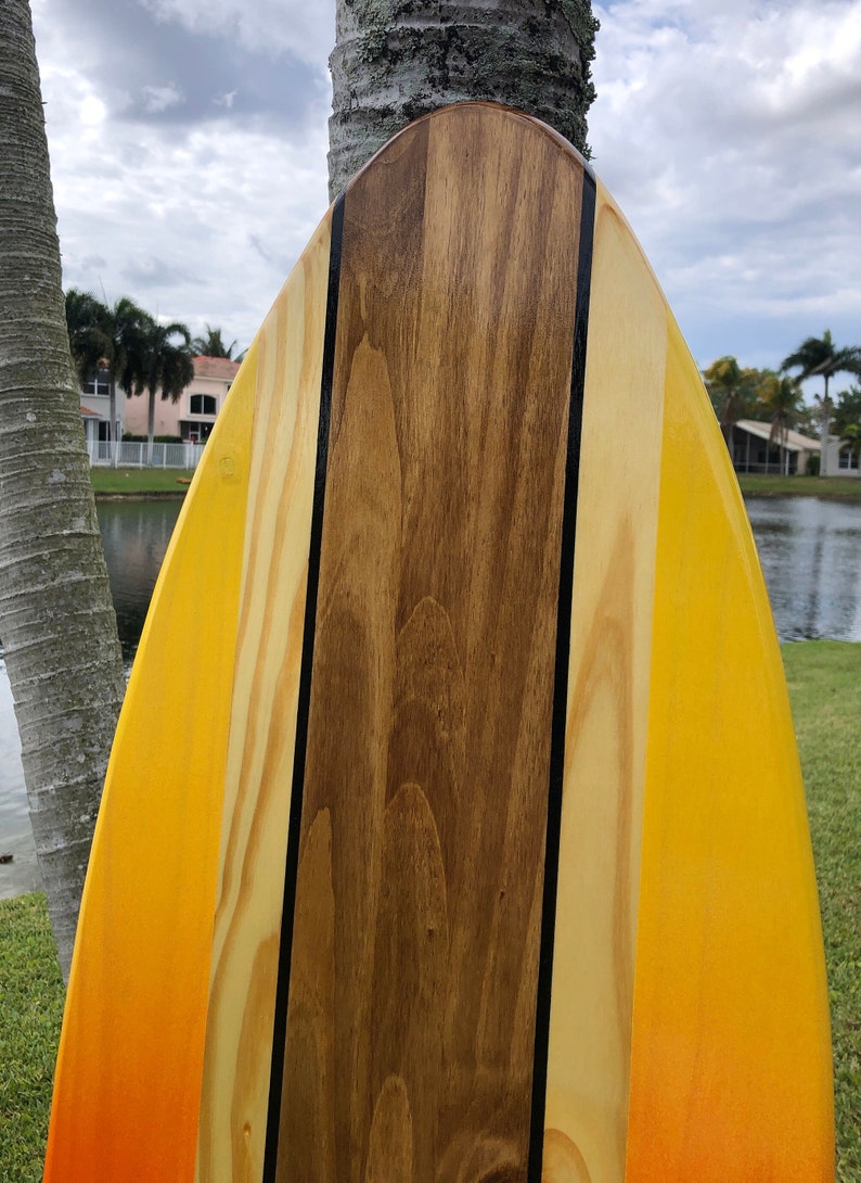 May include: A wooden surfboard with a yellow and orange gradient paint job. The surfboard has a black stripe down the center and a brown wood panel in the middle.