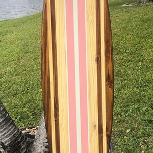 Puede incluir: Una tabla de surf de madera con rayas rosas y blancas pintadas en la superficie. La tabla de surf est&aacute; hecha de diferentes tipos de madera, creando un patr&oacute;n &uacute;nico.