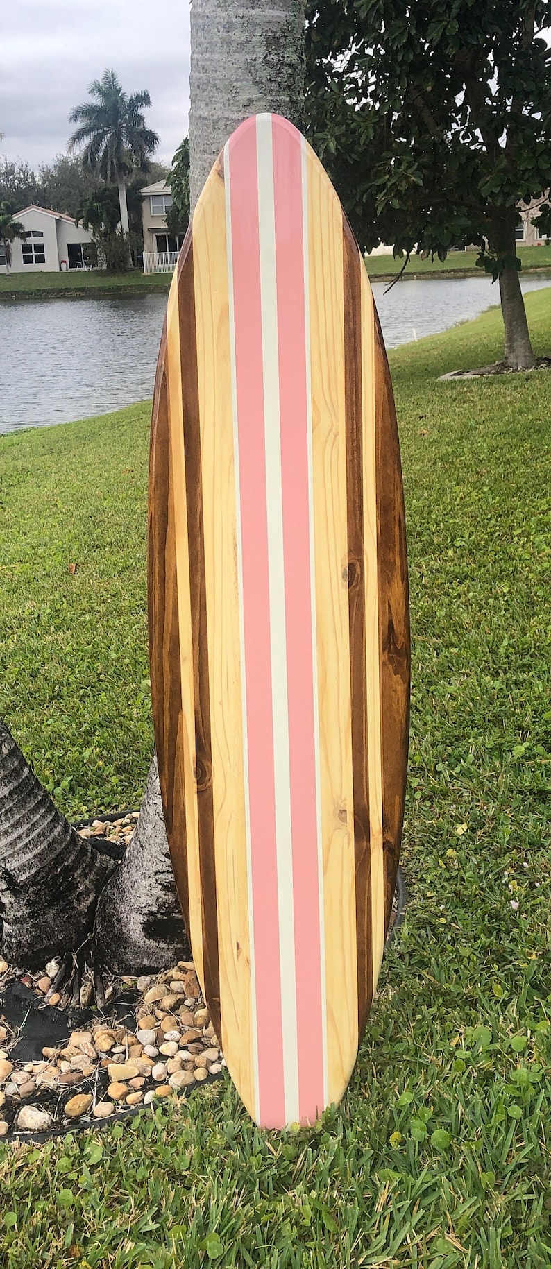 Puede incluir: Una tabla de surf de madera con rayas rosas y blancas pintadas en la superficie. La tabla de surf est&aacute; hecha de diferentes tipos de madera, creando un patr&oacute;n &uacute;nico.