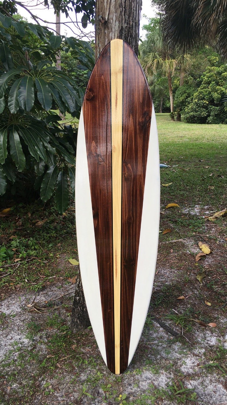 K&ouml;nnte beinhalten: Ein wei&szlig;es Surfbrett mit einem braunen und gelben Holzmaserungsmuster. Das Surfbrett lehnt an einem Baumstamm.