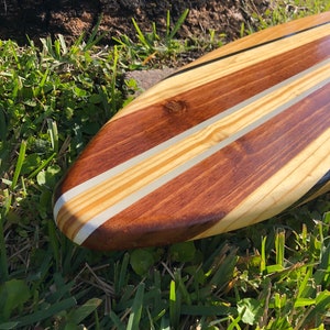 Op de afbeelding: Een houten surfplank met een natuurlijke houten afwerking en zwarte en witte strepen. De surfplank ligt op groen gras.