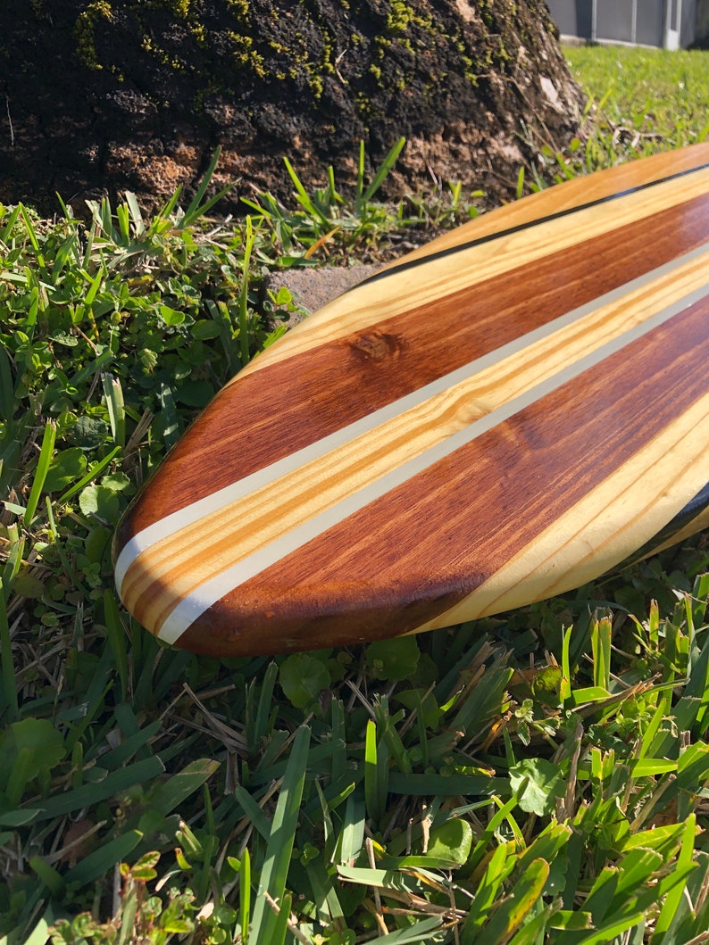 Op de afbeelding: Een houten surfplank met een natuurlijke houten afwerking en zwarte en witte strepen. De surfplank ligt op groen gras.