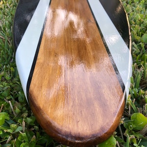 May include: A wooden paddle with a glossy finish. The paddle features a central brown section with black and white diagonal stripes on the edges. The paddle is resting on a bed of green grass.