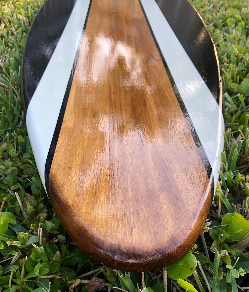 May include: A wooden paddle with a glossy finish. The paddle features a central brown section with black and white diagonal stripes on the edges. The paddle is resting on a bed of green grass.
