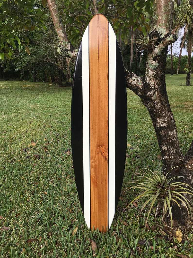 May include: A wooden surfboard with a black, white, and brown design. The surfboard has a long, oval shape with a black border, a central brown stripe, and two white stripes. The board is displayed upright on a grassy surface.