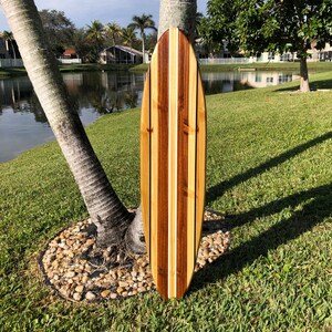 Op de afbeelding: Een houten surfboard met een gestreept ontwerp in lichtbruin, donkerbruin en wit. Het surfboard leunt tegen een palmboom.