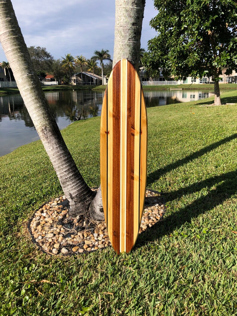 Op de afbeelding: Een houten surfboard met een gestreept ontwerp in lichtbruin, donkerbruin en wit. Het surfboard leunt tegen een palmboom.