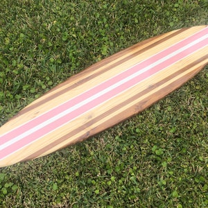 Puede incluir: Una tabla de surf de madera con rayas rosas pintadas en ella. La tabla de surf est&aacute; tumbada sobre un lecho de hierba verde.