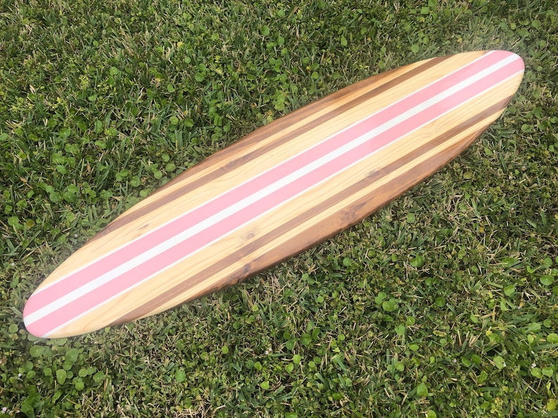 Puede incluir: Una tabla de surf de madera con rayas rosas pintadas en ella. La tabla de surf est&aacute; tumbada sobre un lecho de hierba verde.
