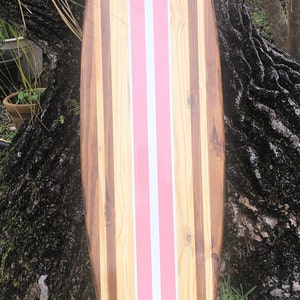 Puede incluir: Una tabla de surf de madera con rayas rosas y blancas. La tabla de surf est&aacute; hecha de diferentes tipos de madera, creando un patr&oacute;n &uacute;nico.