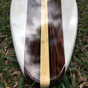 K&ouml;nnte beinhalten: Ein wei&szlig;es Surfbrett mit einem dunklen Holz- und hellem Holzeinlege-Design. Das Surfbrett liegt auf einem Bett aus gr&uuml;nem Gras.