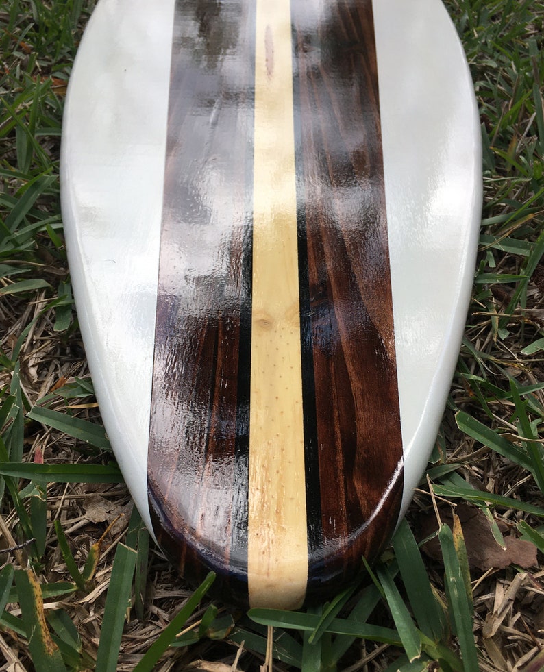 K&ouml;nnte beinhalten: Ein wei&szlig;es Surfbrett mit einem dunklen Holz- und hellem Holzeinlege-Design. Das Surfbrett liegt auf einem Bett aus gr&uuml;nem Gras.