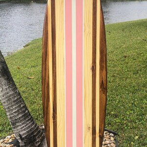 Puede incluir: Una tabla de surf de madera con un dise&ntilde;o de rayas rosas y blancas. La tabla de surf est&aacute; hecha de diferentes tipos de madera, incluyendo madera marr&oacute;n y madera clara.