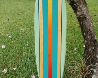 May include: A teal and yellow surfboard with a red stripe down the center. The surfboard is leaning against a tree trunk in a grassy area.