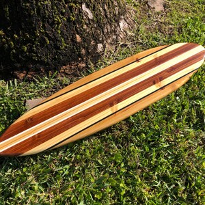 Op de afbeelding: Een houten surfboard met een natuurlijke houten afwerking en twee witte strepen. Het surfboard ligt op een bed van groen gras.