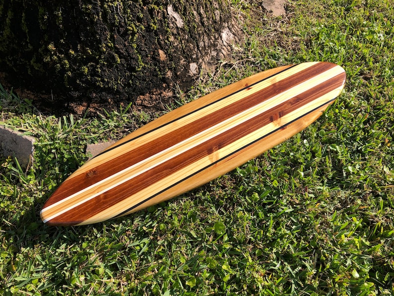 Op de afbeelding: Een houten surfboard met een natuurlijke houten afwerking en twee witte strepen. Het surfboard ligt op een bed van groen gras.