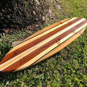Op de afbeelding: Een houten surfboard met een natuurlijke houten afwerking. Het surfboard heeft drie witte strepen en zwarte accenten.