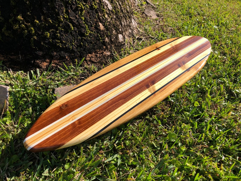 Op de afbeelding: Een houten surfboard met een natuurlijke houten afwerking. Het surfboard heeft drie witte strepen en zwarte accenten.