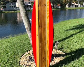 May include: A red and brown wooden surfboard with a striped design. The surfboard is leaning against a palm tree in a grassy area.